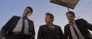 Mr. Blonde, Mr. White, and Mr. Pink look into the trunk with the tied-up officer inside.
